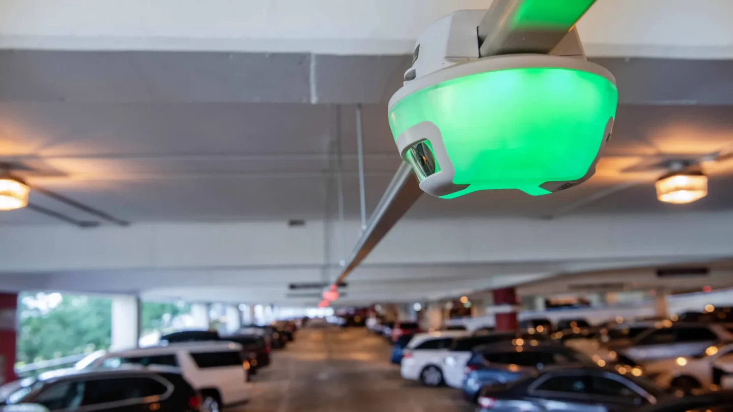 A close-up of a green M5 smart sensor, the heart of Park Assist's parking guidance system, installed on the ceiling of a parking garage