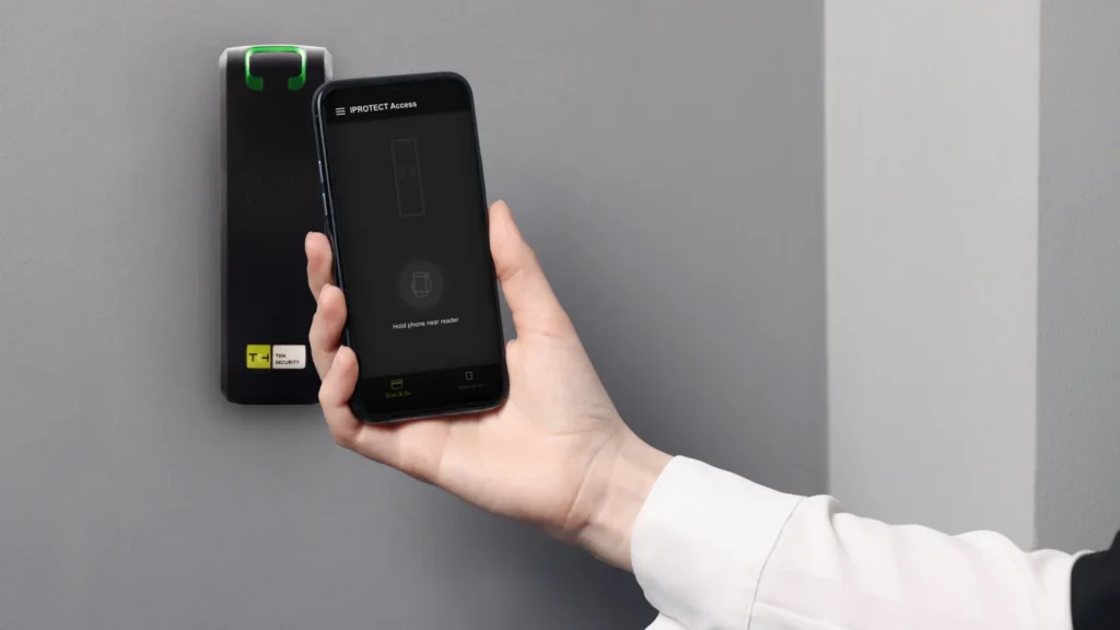 Hand holding a smartphone near a wall-mounted IPROTECT access control reader