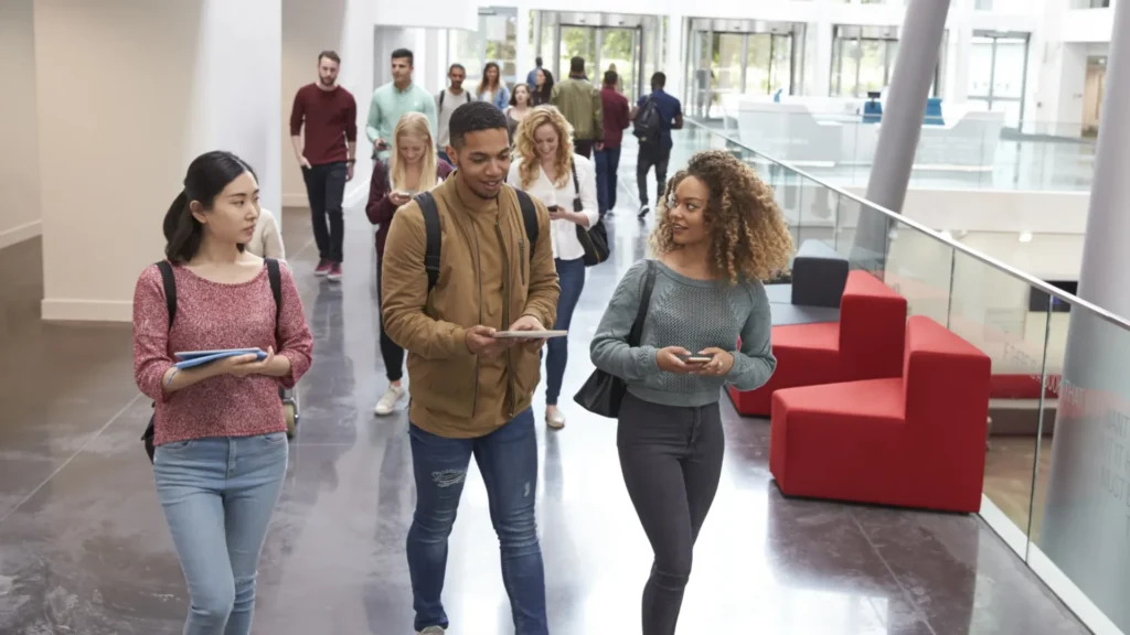 Students walking through a modern building corridor while using smartphones and tablets in Education Sector