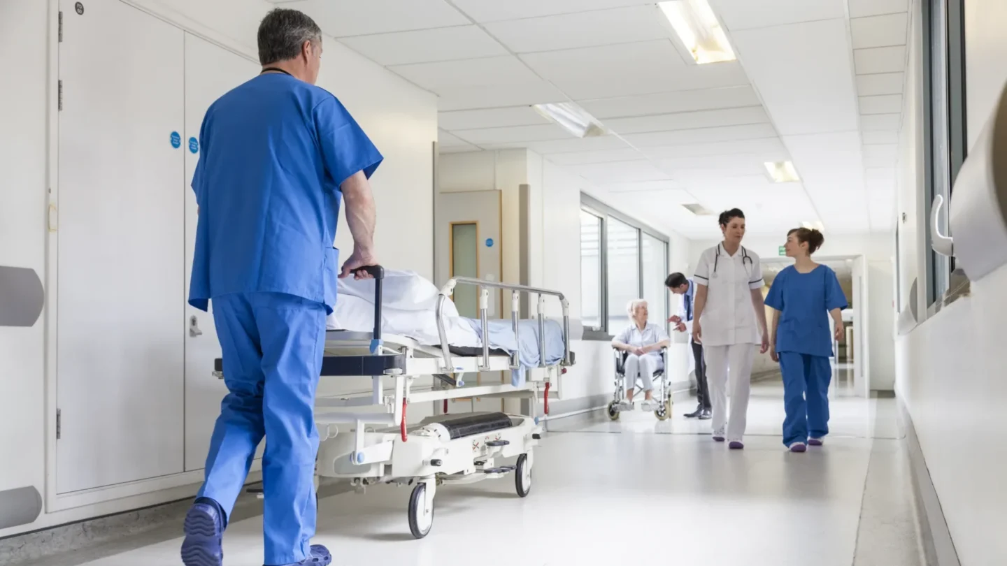 Hospital staff in healthcare sector moving a patient bed through a corridor while other staff walk nearby