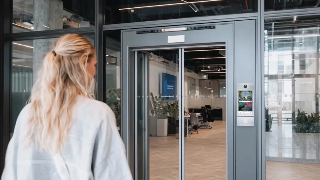 Woman walking toward a glass elevator lobby with a Commend intercom panel in a commercial building