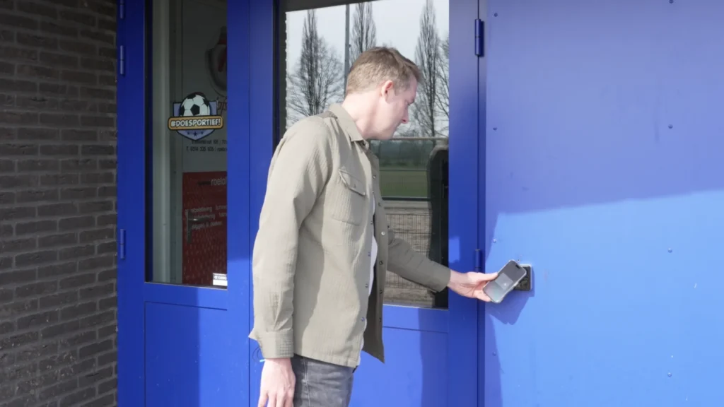 Angerlo Vooruit member using a smartphone to unlock a blue entrance door with the ATLAS mobile access control app.
